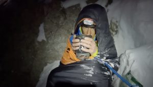 a climber in a sleeping bag, sitting in the snow in the dark